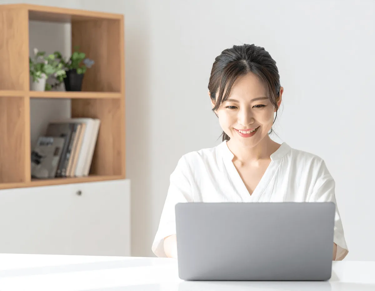 「日本人女性が笑顔でノートパソコンを操作している。自然光が差し込む室内で、背景はシンプル。落ち着いた色合いでやさしい印象」の生成画像