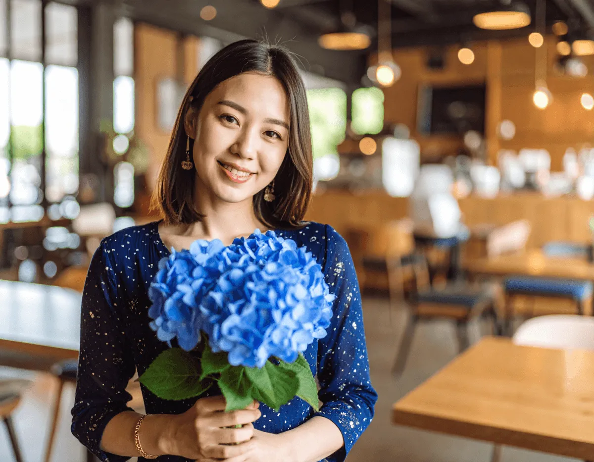「日本人女性がブルーの花束をもってこちらを見ている。背景はカフェの店内。落ち着いた雰囲気。ワイドショット」の生成画像